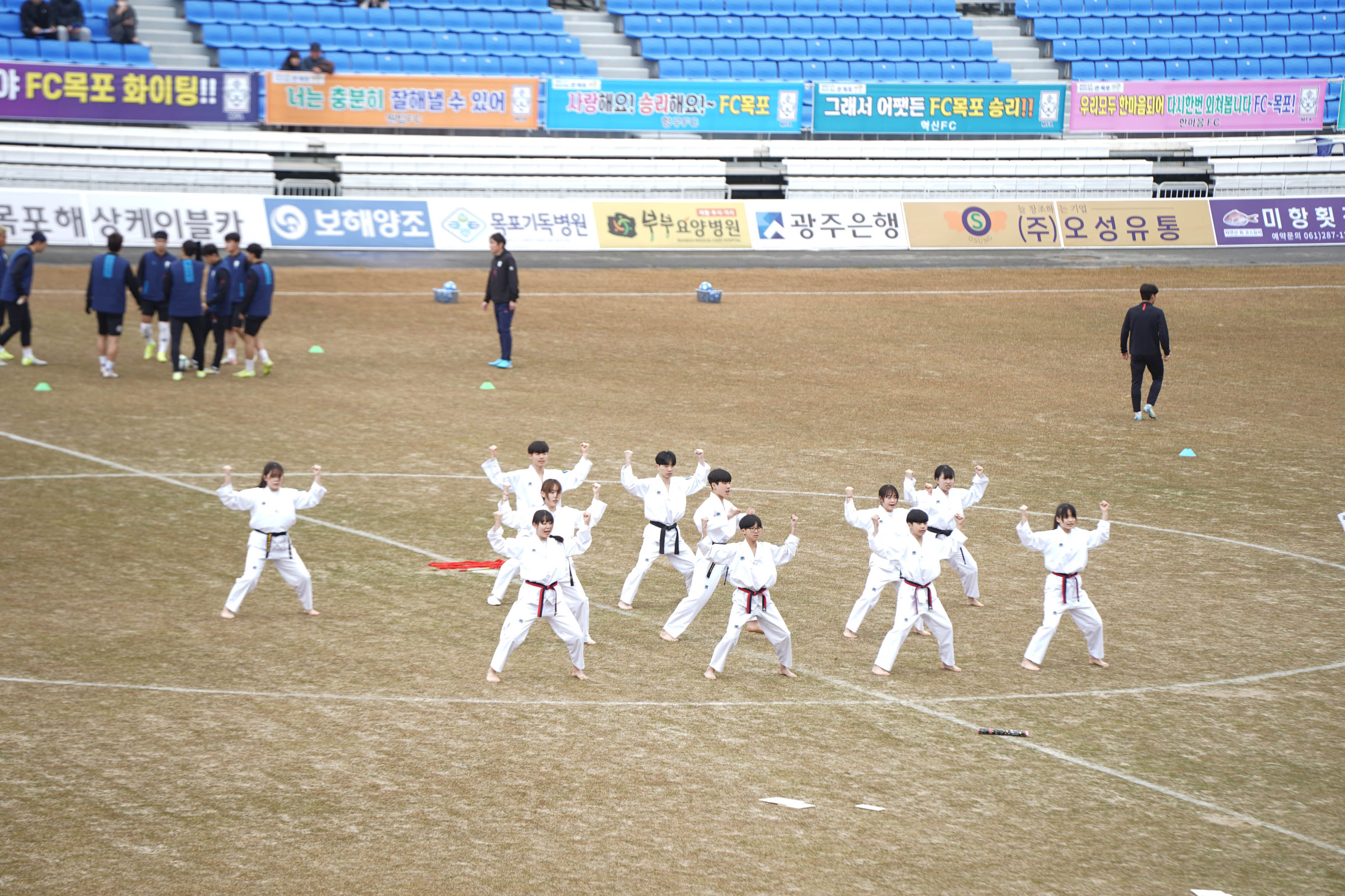 축구 경기장 중앙에서 흰색 태권도 도복을 입은 남녀 학생들이 단체로 태권도 시범을 보이고 있다. 이들은 발을 벌리고 두 팔을 들어 올린 자세로 단체 동작을 선보이고 있으며, 몇몇은 빨간색 또는 검은색 띠를 착용했다. 뒷편 잔디 구역에는 축구 유니폼을 입은 선수들이 준비 운동을 하고 있으며, 관중석에는 '야 FC목포 화이팅!!', '너는 충분히 잘해낼 수 있어', '사랑해요! 승리해요~ FC목포', '그래서 어쨌든 FC목포 승리!!', '우리모두 아낌없이 대신함께 외쳐봅시다 FC목포!!' 등의 다양한 응원 문구가 적힌 현수막이 설치되어 있다. 경기장 광고판에는 '보해양조', '목포기독병원', '부부요양병원', '광주은행', '(주)오성유통', '미항회' 등이 표기되어 있다.