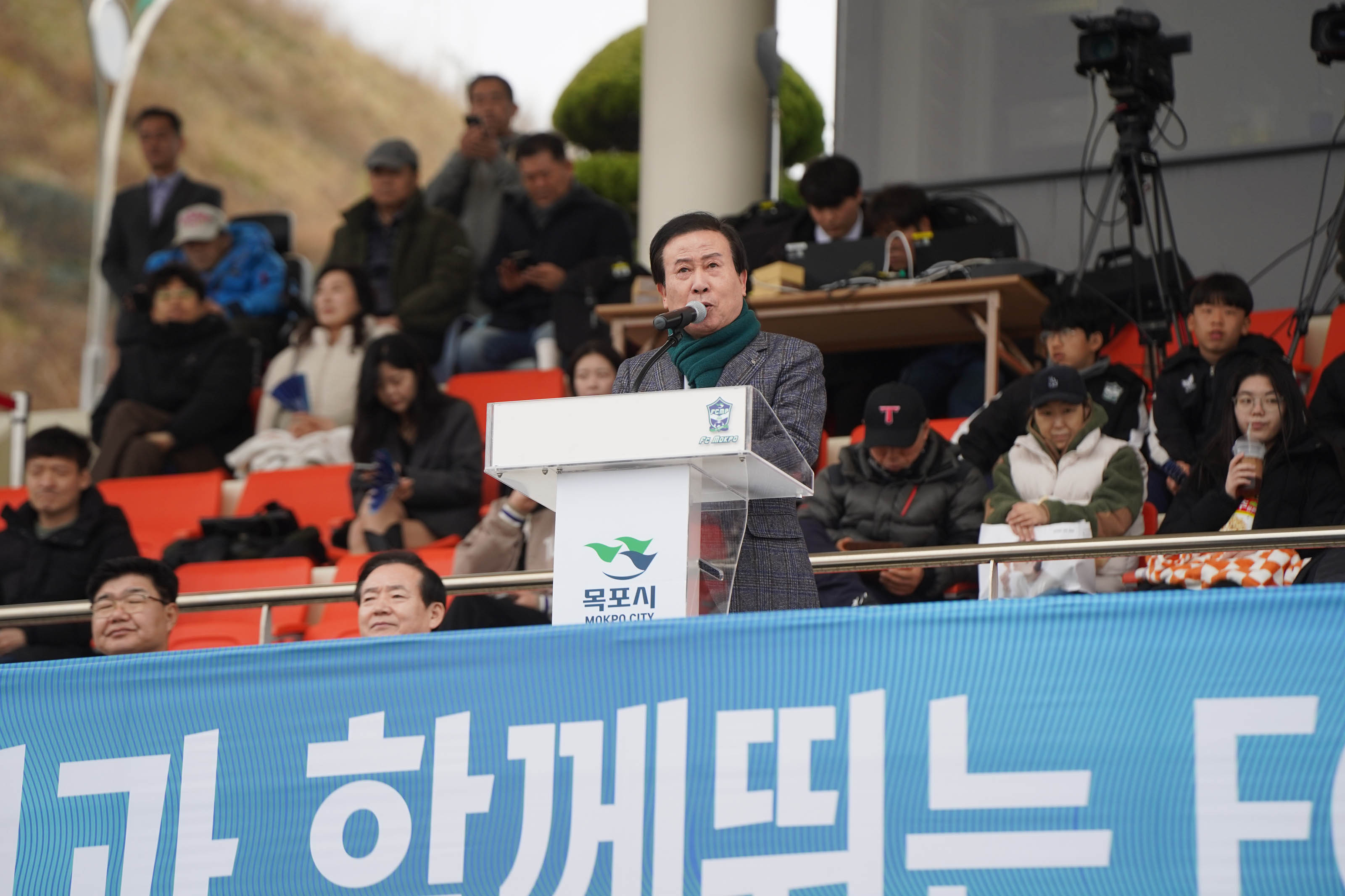 축구장 단상에서 박홍률 목포시장이 '목포시 MOKPO CITY' 로고가 붙은 연단 앞에서 마이크를 통해 개회사를 하고 있다. 뒤쪽 좌석에는 일반 시민들과 학생들이 착석해 있으며, 몇몇은 휴대폰으로 사진을 찍거나 촬영 중이다. 연단 앞에는 파란색 배너가 설치되어 있고, 그 위에는 '시민과 함께 뛰는 FC목포' 라는 문구가 적혀 있다.