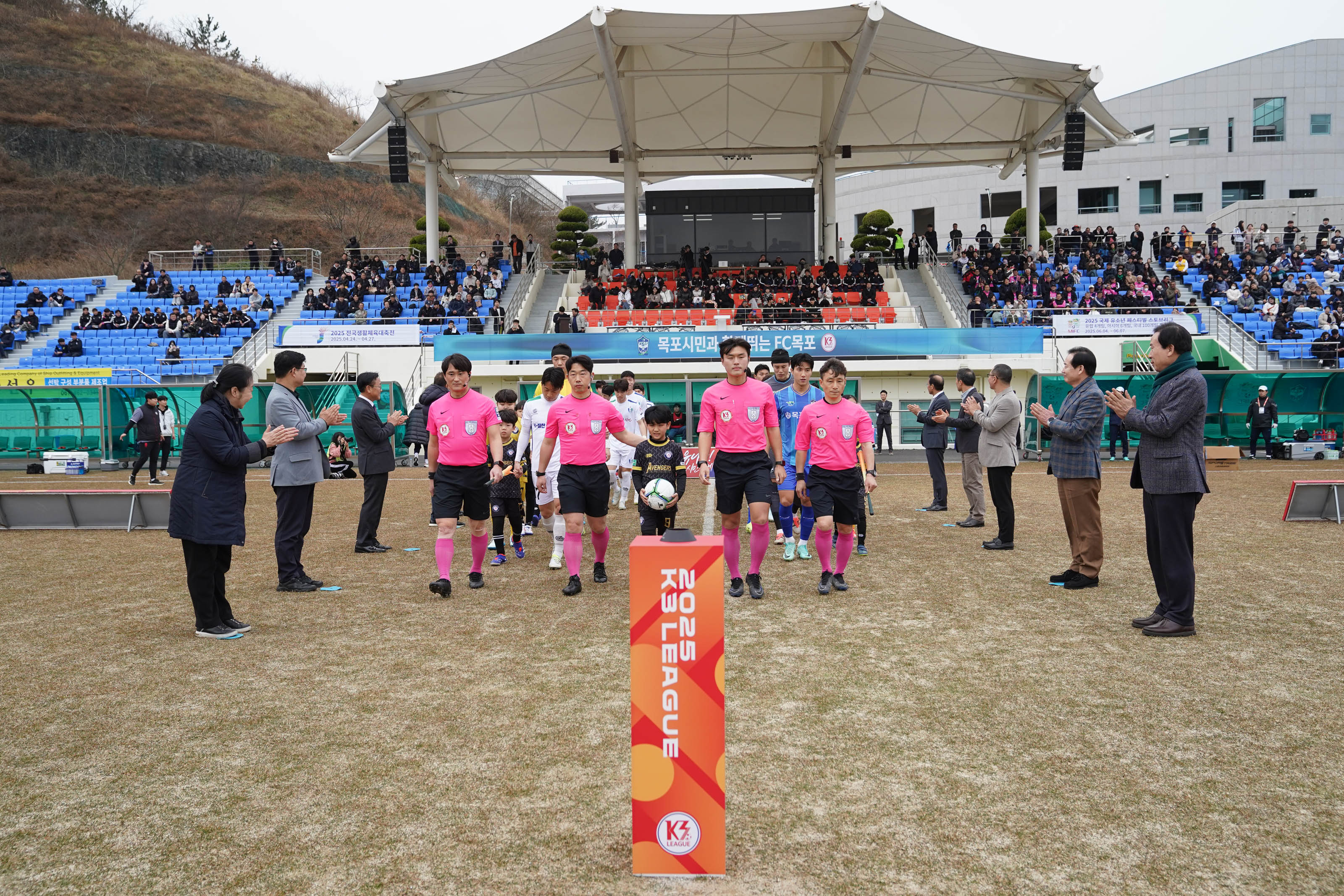 '2025 K3 LEAGUE' 라고 적힌 기둥을 중심으로 심판진과 축구 선수들이 경기장 입장식을 진행하고 있다. 맨 앞의 선수들은 분홍색 유니폼을 입었고, 선수들은 흰색과 파란색 유니폼을 착용했다. 경기장 양쪽에는 박수를 치며 환영하는 내빈들이 도열해 있으며, 관중석은 파란색 좌석으로 채워져 있다. 중앙에는 '목포시민과 함께 뛰는 FC목포'라는 문구가 적힌 현수막이 걸려 있다.