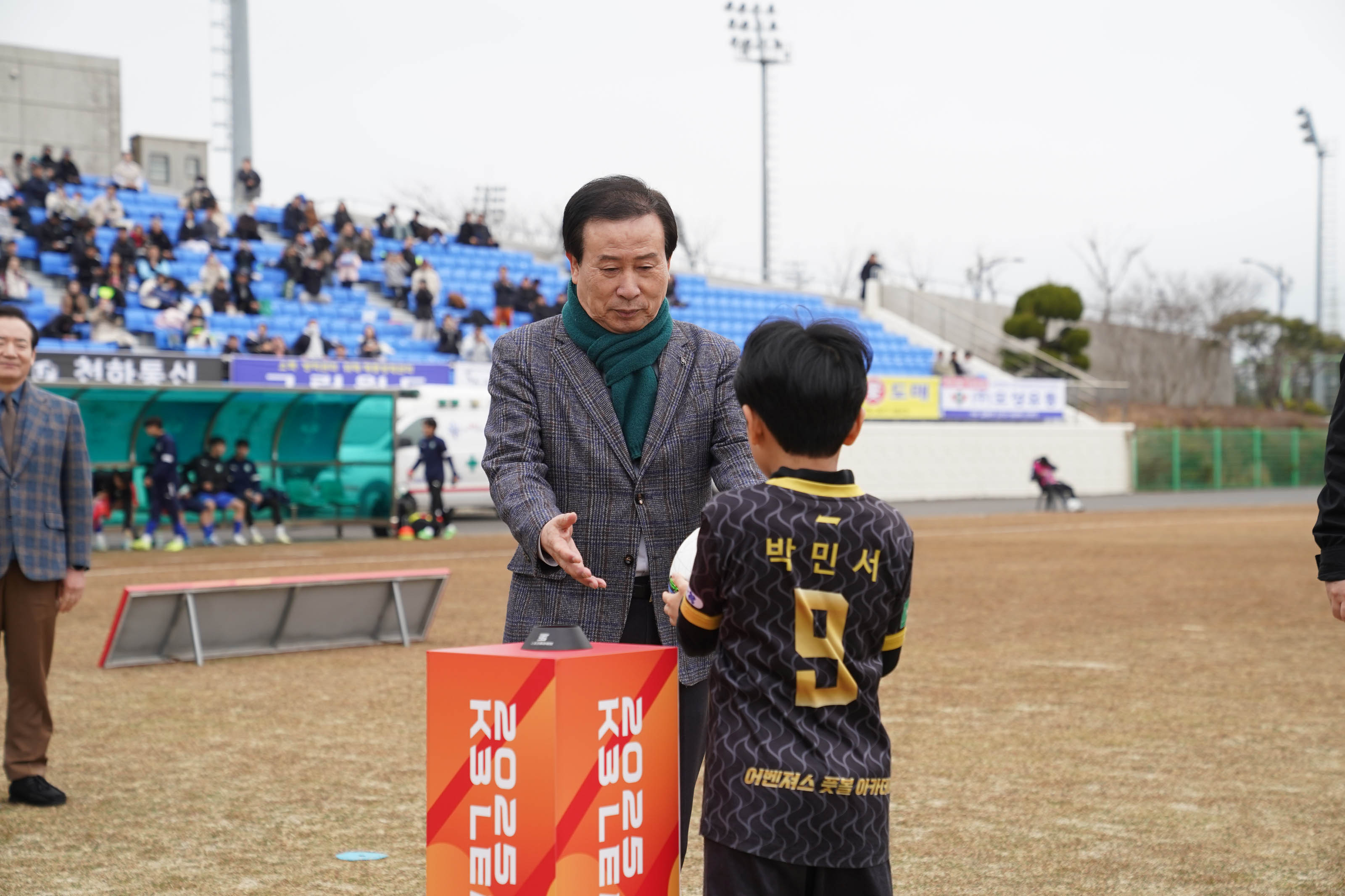 '2025 K3 LEAGUE' 기둥 앞에서 박홍률 목포시장이 축구공을 들고 있는 어린이 선수에게 다가가고 있다. 아이의 유니폼 뒷면에는 '박민서'와 '어벤져스 축구 아카데미' 라는 글자가 새겨져 있다. 뒤편에는 관중들이 앉아 있는 파란색 좌석과 광고판이 있으며, 경기장 옆에는 벤치에 앉아 있는 선수들이 보인다.