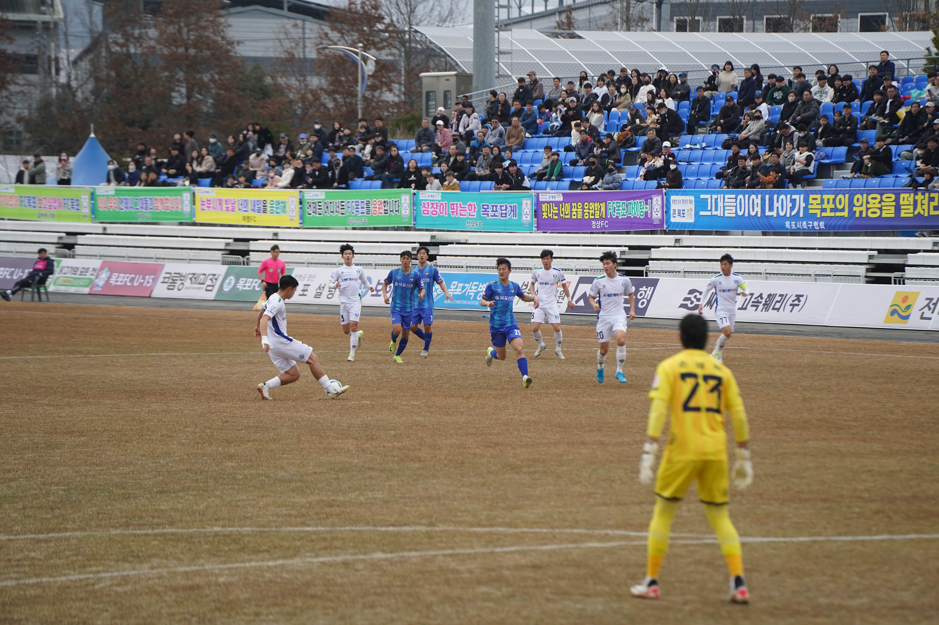 축구 경기장에서 흰색 유니폼을 입은 선수가 왼발로 롱패스를 시도하는 장면. 상대팀 파란색 유니폼의 선수들이 달려오고 있으며, 경기장은 많은 관중으로 가득 찼다. 배경에는 '그대들이여 나아가 목포의 위용을 펼쳐라 FC목포 파이팅!', '심장이 뛰는 한 목포답게', '밝아오는 너의 꿈을 응원할게 FC목포 파이팅!' 등 다양한 응원 문구가 적힌 현수막들이 스탠드에 설치되어 있다.