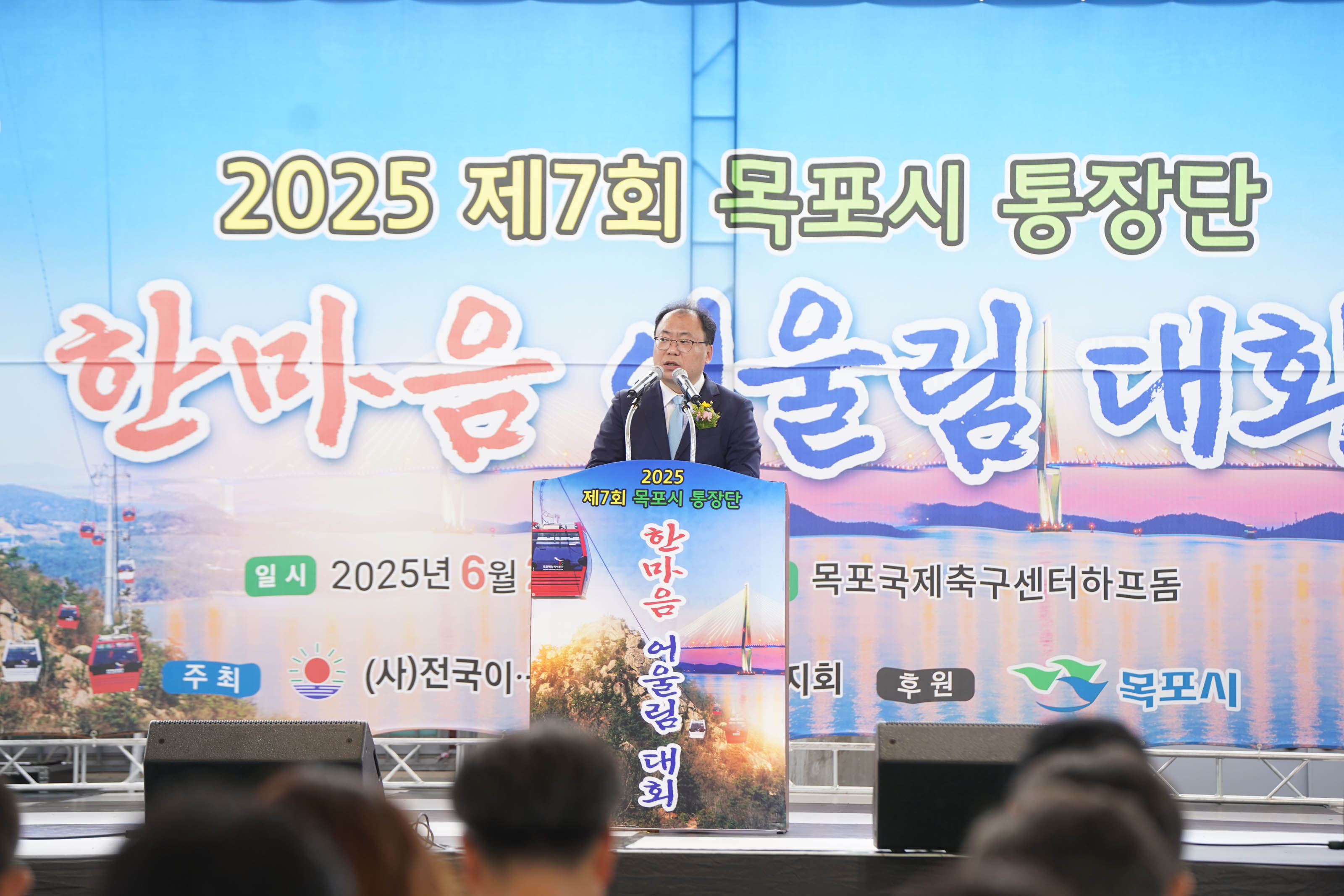 2025 제7회 목포시 통장단 한마음 어울림 대회 무대 위의 연설자의 모습. 배경 현수막에는 '희망찬 도약! 청년이 찾는 큰 목포' 문구와 유달산 케이블카, 목포대교 이미지가 그려져 있다. 일시는 2025년 6월 20일(금), 장소는 목포국제축구센터 하프돔이다.