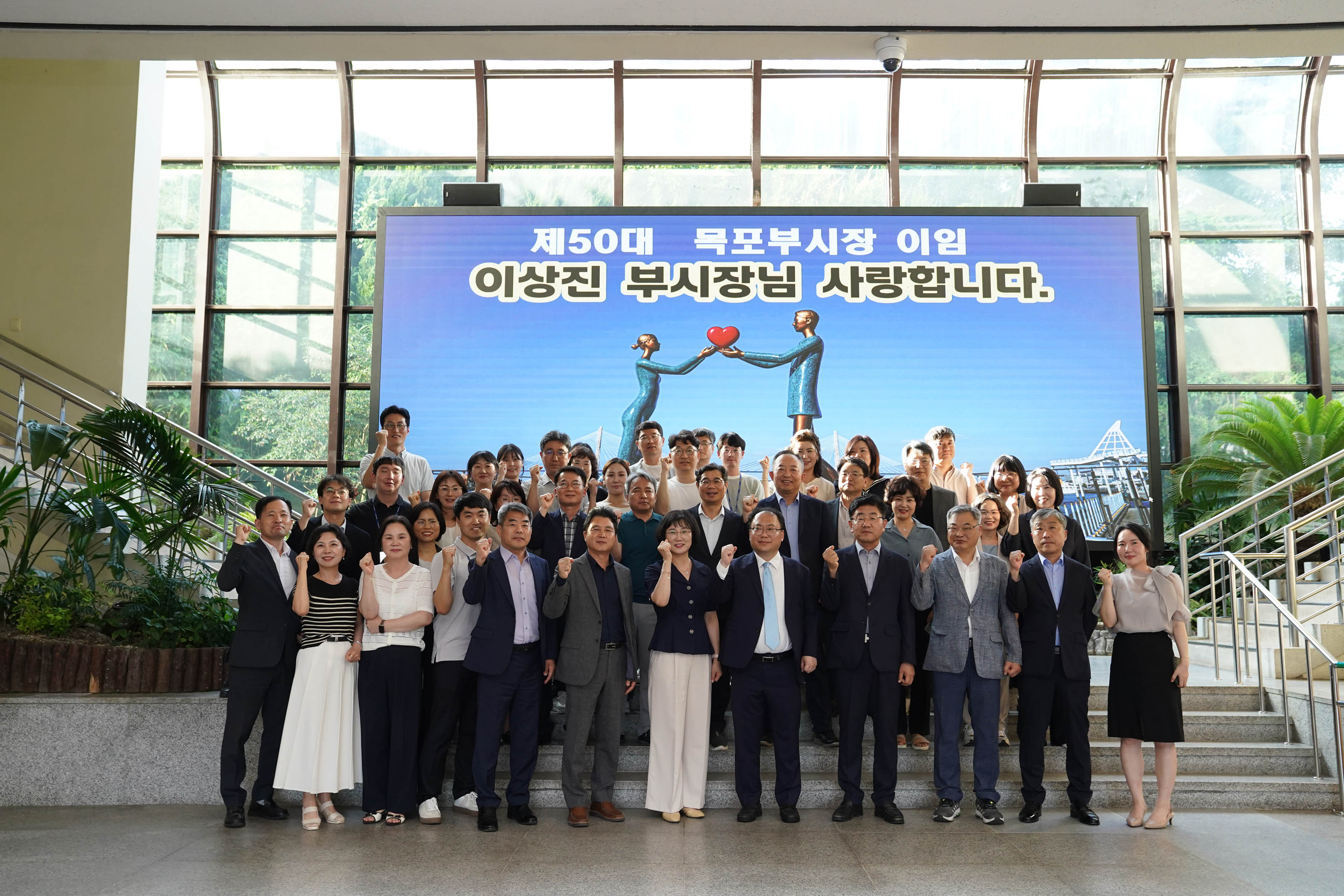 '제50대 목포부시장 이임 이상진 부시장님 사랑합니다'라는 문구가 적힌 대형 스크린 앞에서 수십 명의 목포시청 직원들이 단체로 모여 주먹을 쥐고 파이팅 포즈를 취하며 기념 촬영을 하는 모습.