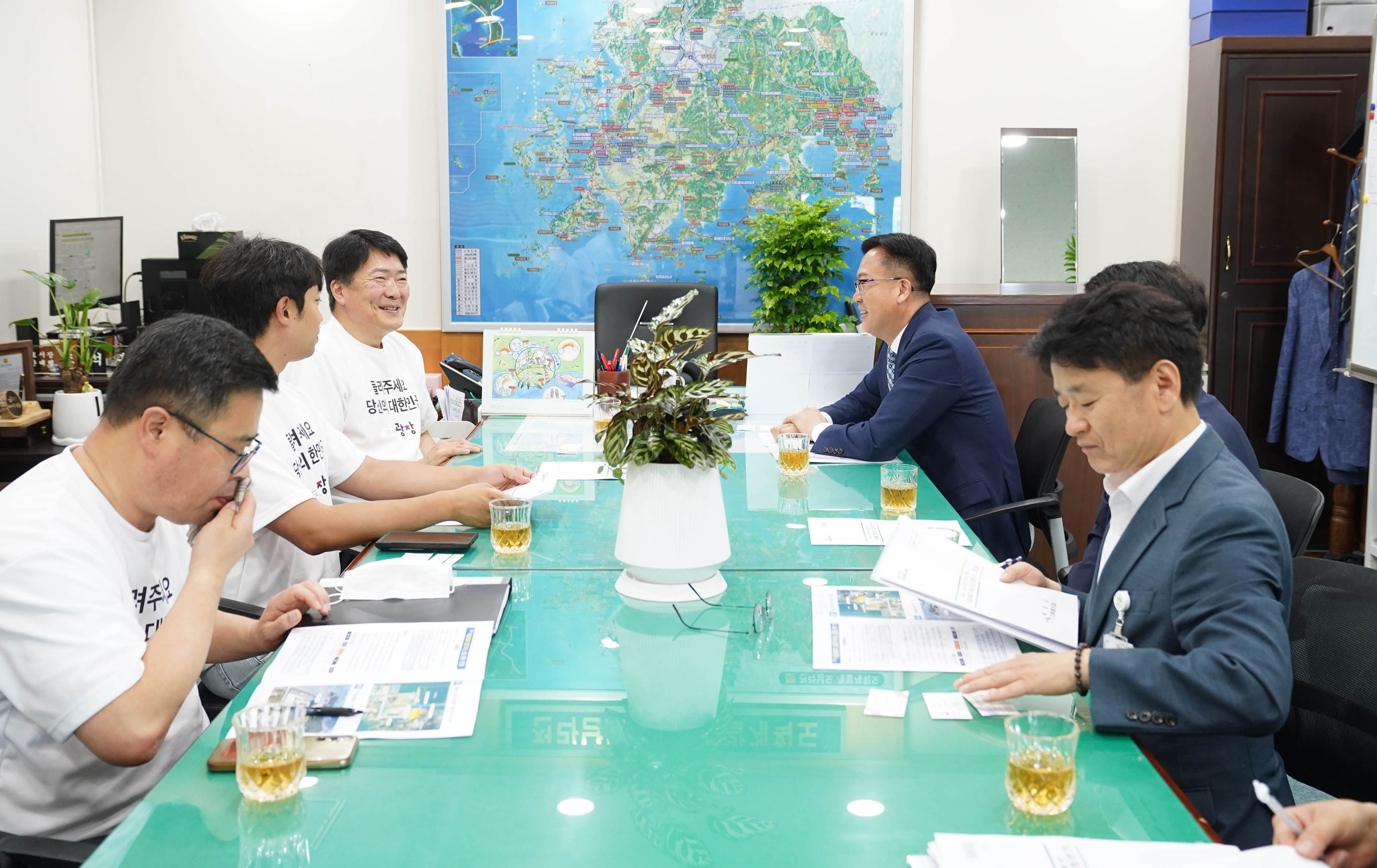 녹색 회의 테이블에 앉은 참석자들이 서류를 읽거나 대화를 나누는 전체적인 회의 현장 모습.