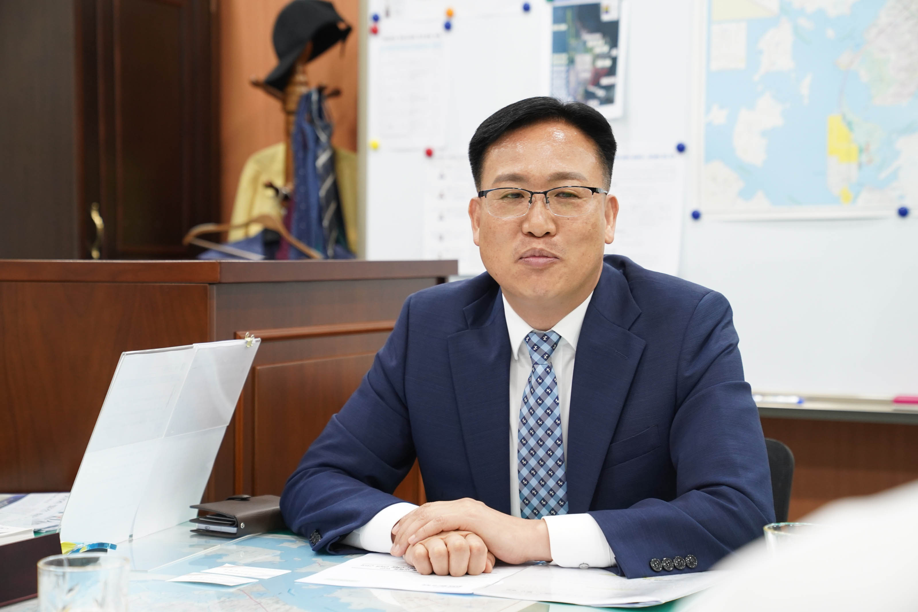 안경을 쓰고 네이비색 정장에 체크무늬 넥타이를 맨 남성이 책상 앞에 앉아 정면을 응시하고 있는 모습. 배경에는 지도가 붙은 화이트보드와 옷걸이가 보임.
