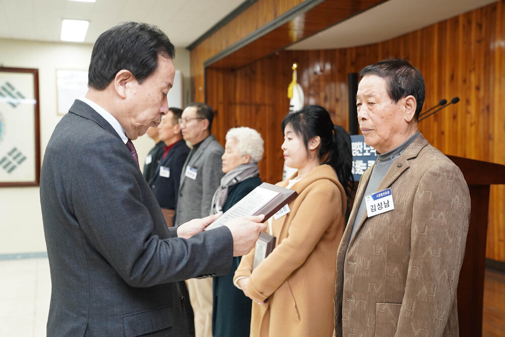 3월 정례조회에서 정장입은 남성이 갈색 자켓을 입은 남성에게 패를 건내고 있다.