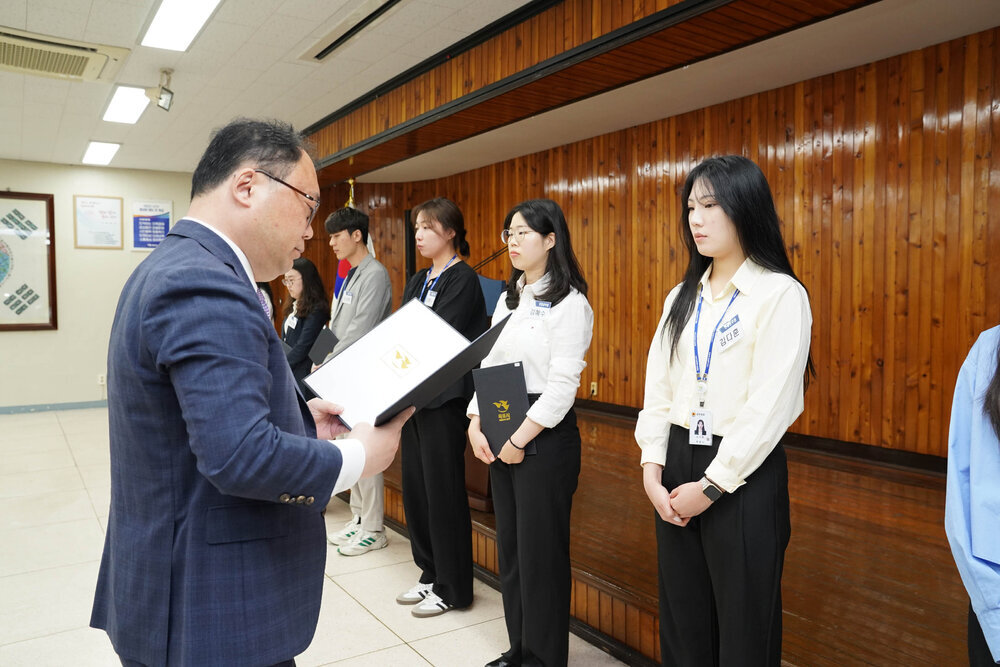 정장을 입고 안경을 낀 남성이 연노랑 셔츠를 입은 여성에게 상장을 수여하고 있다