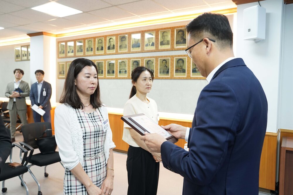 정장 차림의 관계자가 체크 원피스를 입은 여성 수상자에게 표창패를 전달하며 읽고 있는 모습이다.