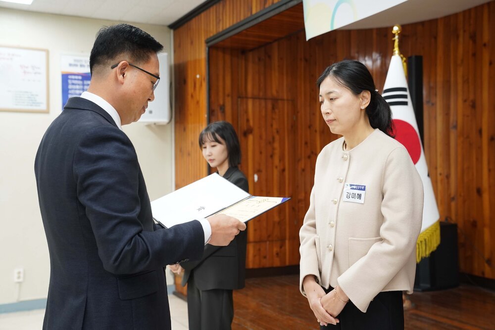 11월 정례조회로 정장을 입고 안경을 쓴 남성이 긴머리를 뒤로 묵고 밝은 베이지색 짧은 코트를 입은 강미혜 수상자에게 상장을 수여 하고 있다.