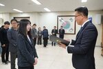 2026년 상반기 4급이하 정기인사로 정장을 입은 남성이 실내 행사장에서 한 여성에게 상장을 전달하며 내용을 읽고 있고, 여성은 남색 트위드 자켓을 입고 사원증을 착용한 채 두 손을 모은 상태로 서 있으며 뒤쪽으로 참석자들이 줄지어 서서 행사를 지켜보고 있다.